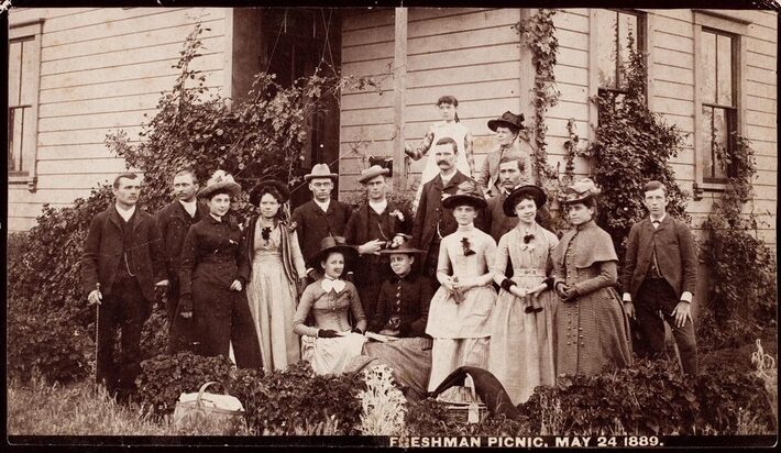 Photograph of USC freshmen picnic, May 24, 1889. A group of seven men and nine women can be seen standing outside a building, surrounded by ivy and other foliage.
