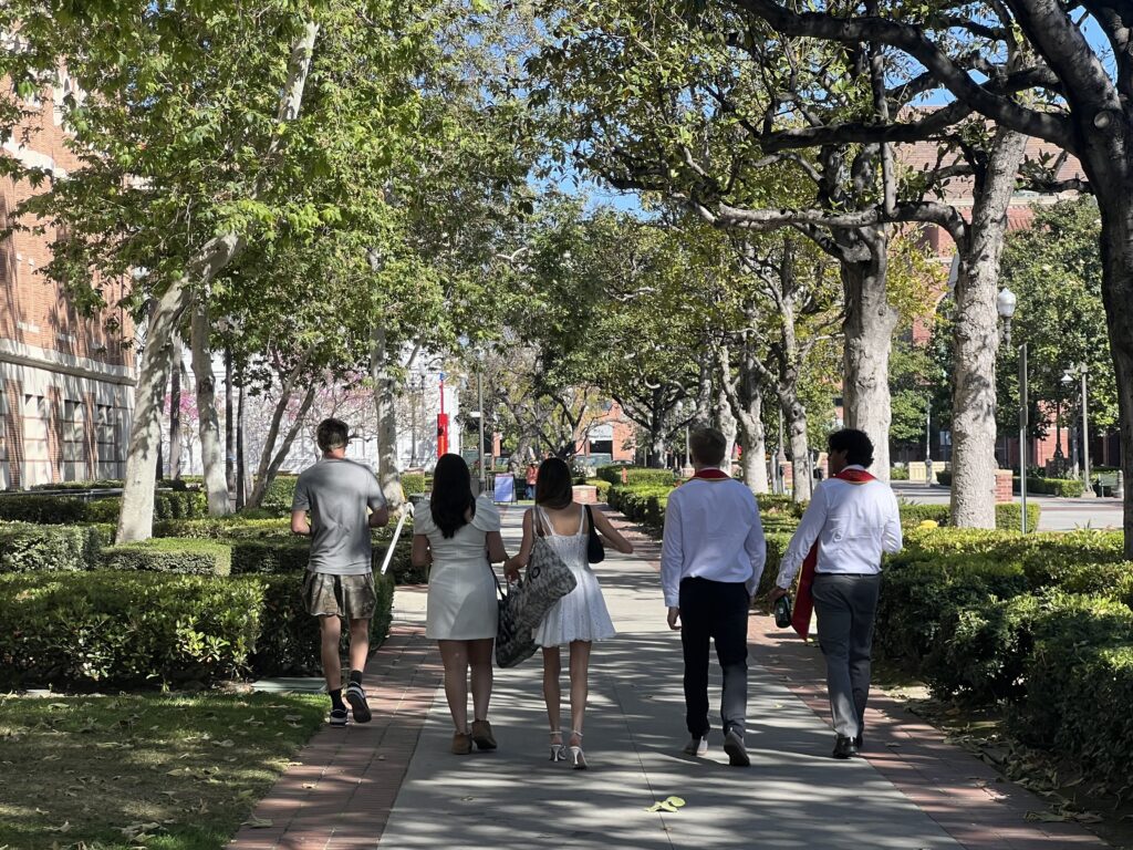 Five USC students walk a tree lined path around Doheny Library