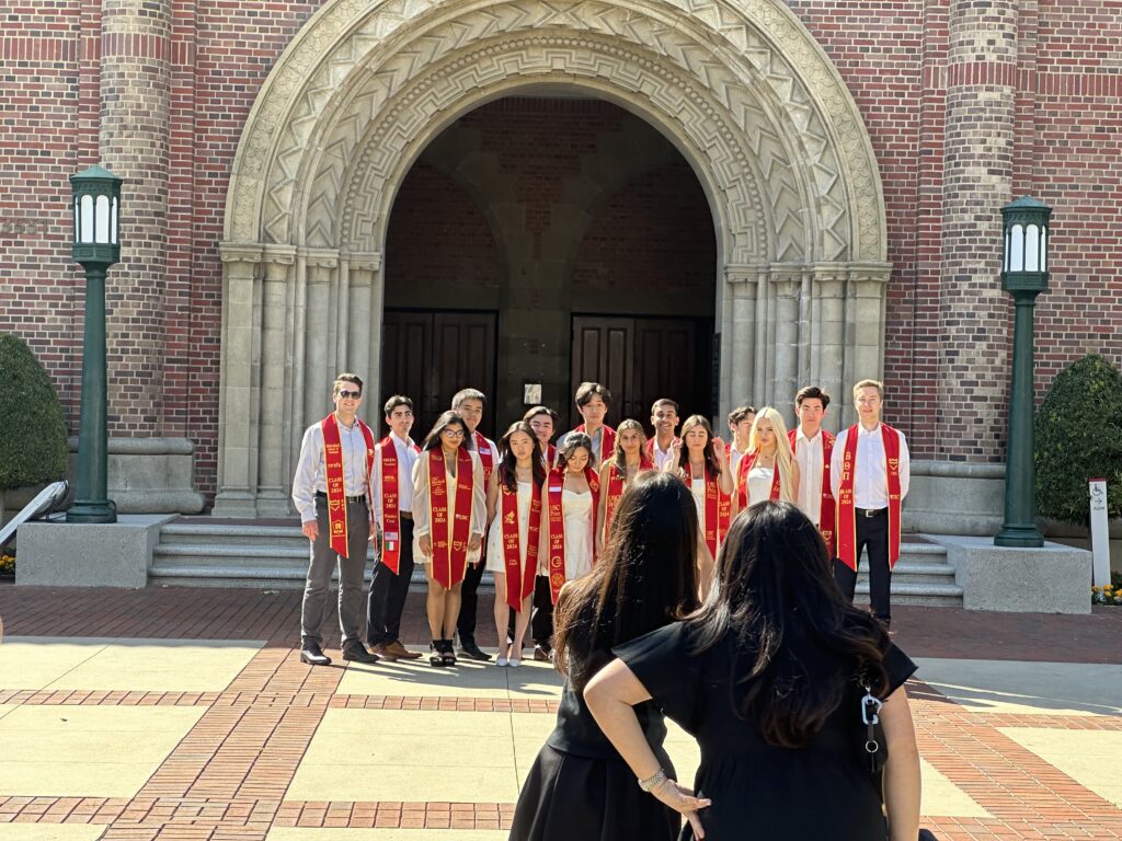 A group of 15 Trojans pose for a group photo with their graduation sashes in front of Bovard Auditorium